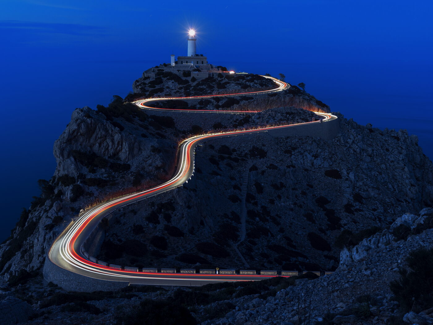 Lighthouse on a headland at blue hour, light trails winding up the road below