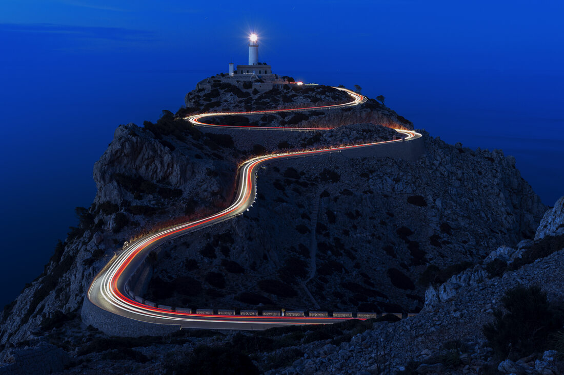 Lighthouse on a headland at blue hour, light trails winding up the road below